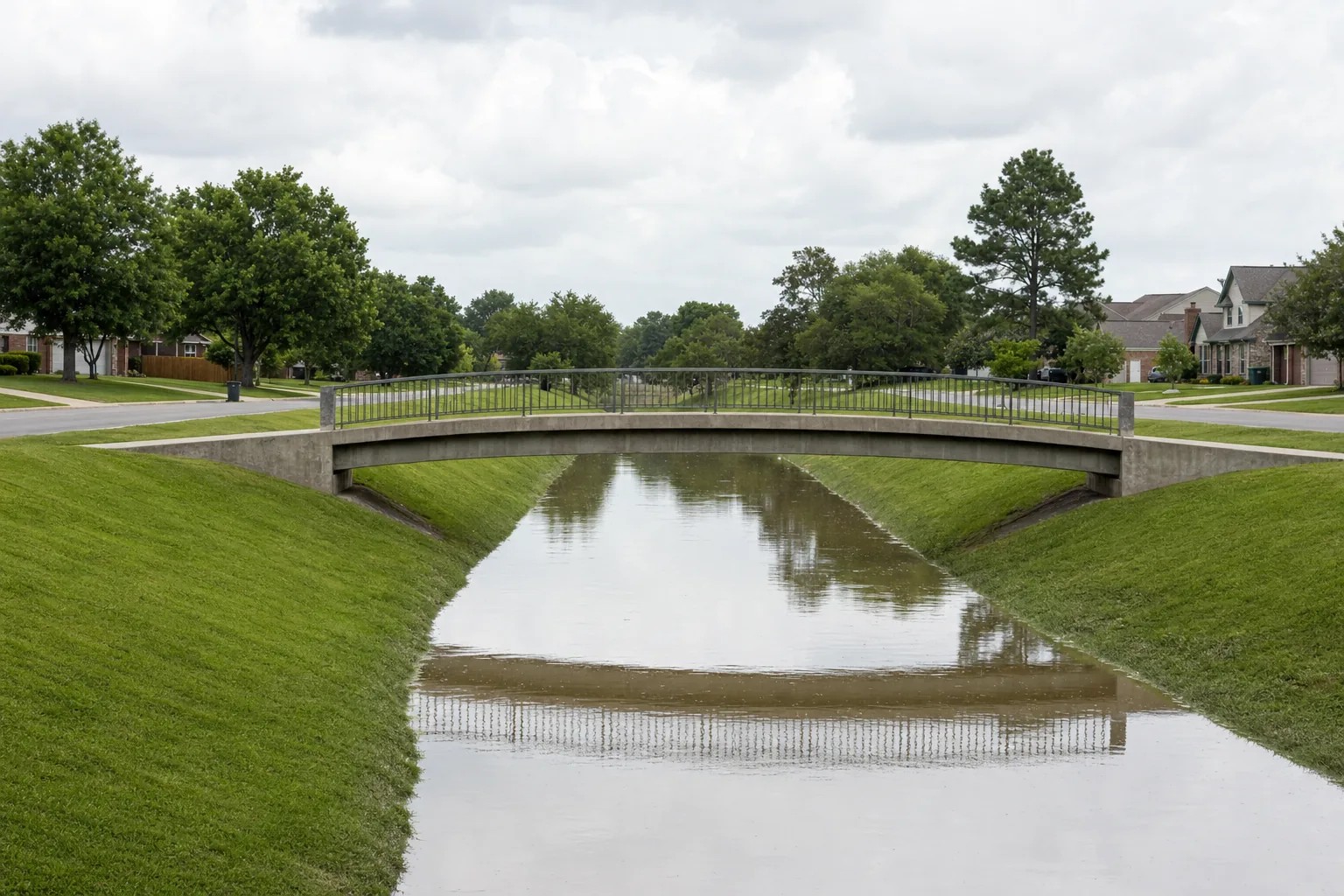 A wide drainage canal with mown grass banks running between streets, crossed by a concrete footbridge